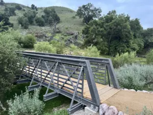 Slate-gray FRP truss bridge with a wood deck crossing a creek in a green, oak-covered canyon.