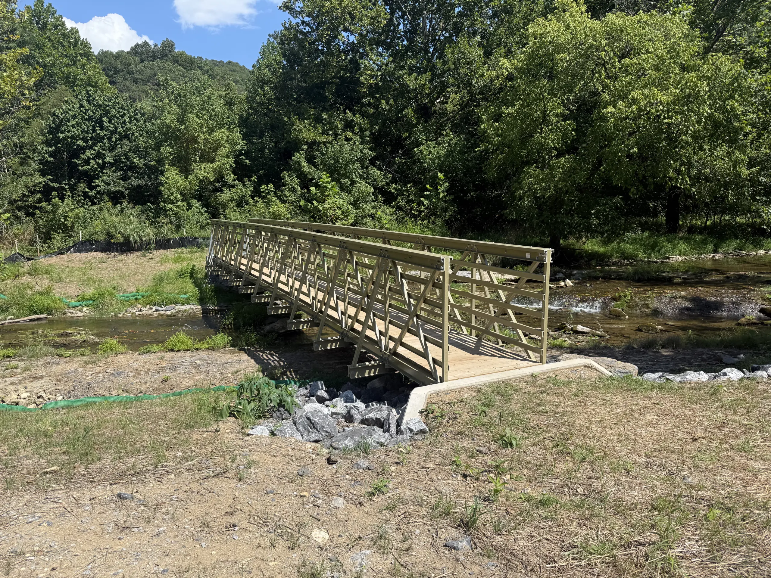 Completed olive-green FRP pedestrian truss bridge with timber decking spanning rocky Catawba Creek on the Andy Layne Trail, framed by a forested hillside.