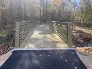 A 60-foot FRP truss trail bridge with wooden decking spanning across a creek along a paved trail surrounded by autumn trees.
