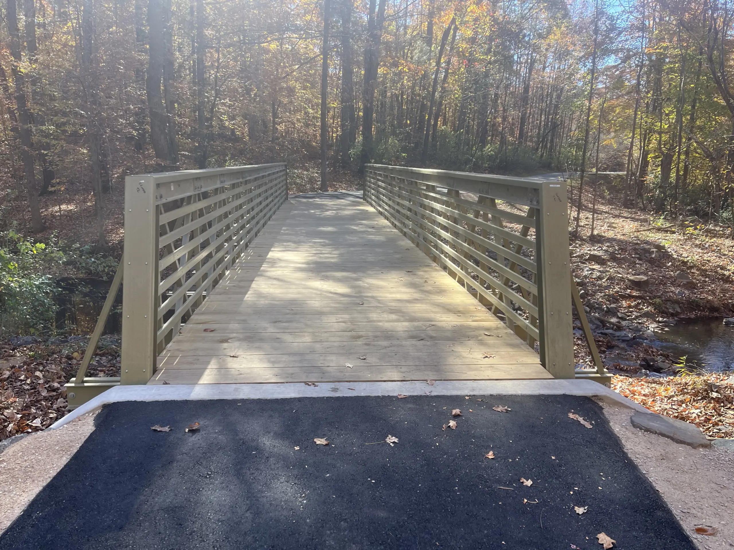 A 60-foot FRP truss trail bridge with wooden decking spanning across a creek along a paved trail surrounded by autumn trees.